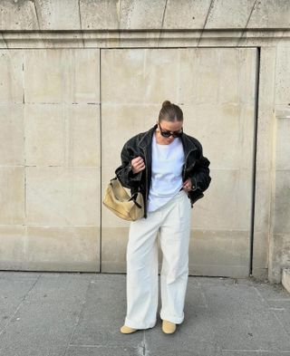 Dina wears ivory wide-leg jeans, a leather jacket, white t-shirt, clogs, and a suede bag.