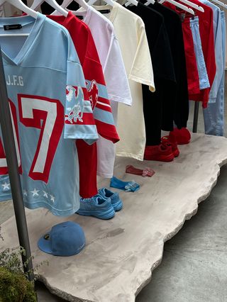 A photo of a rack of clothes, shoes, and accessories from the Joe Freshgoods gallery space during the Paris Olympics called A Friend Named Cousin. Among the pieces are ones from a collaboration with New Balance.