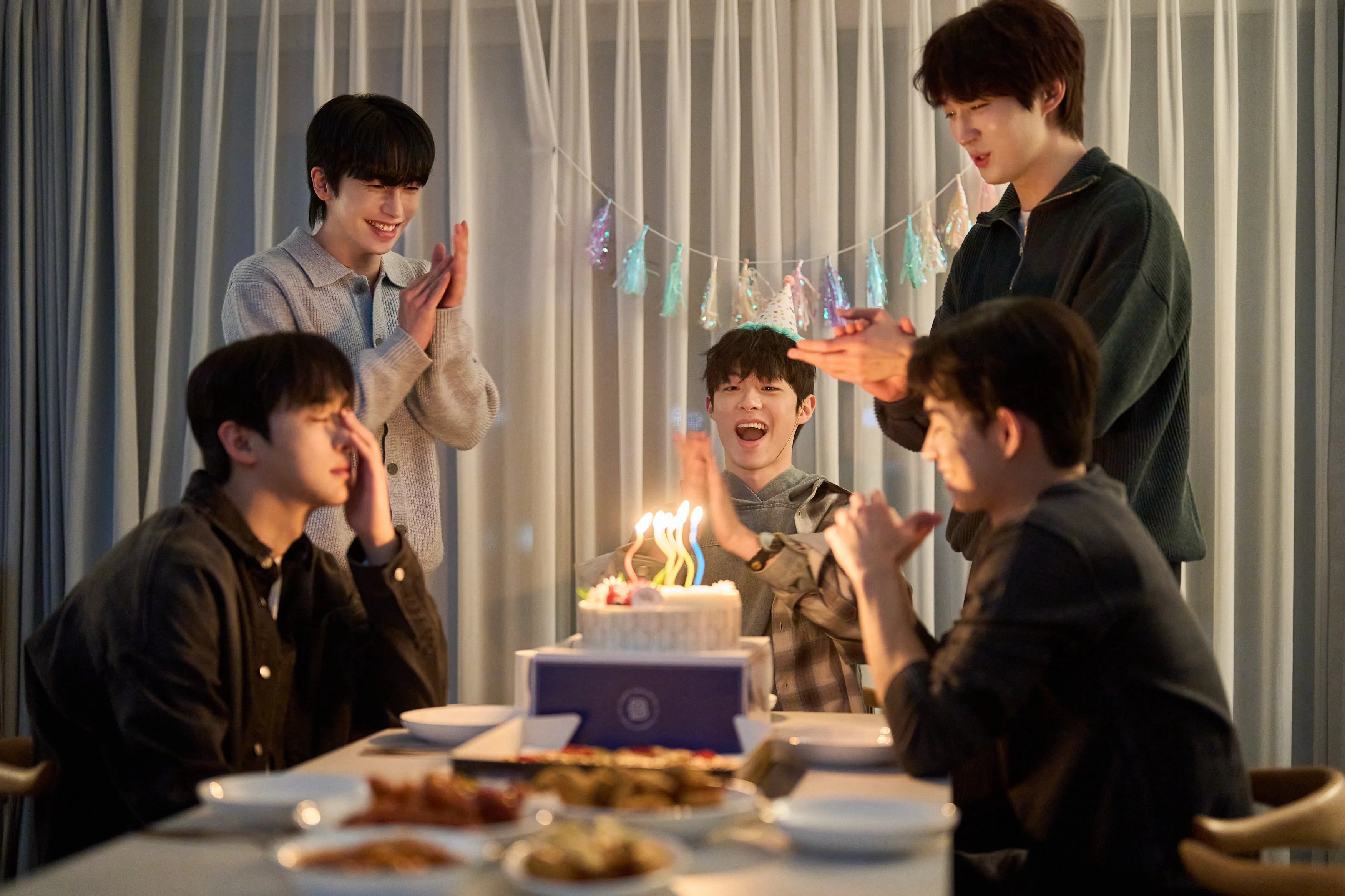 four men sit at a table while one looks upset having to celebrate his birthday and blow out the candles on a cake in a still from the bl kdrama my secret vampire