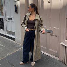 Maya wears black trousers, brown tank, trench coat, and tan adidas. 