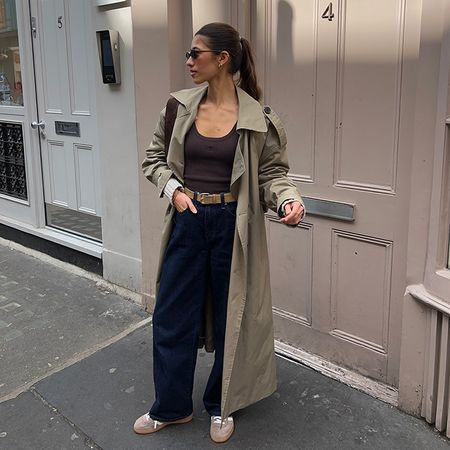 Maya wears black trousers, brown tank, trench coat, and tan adidas.