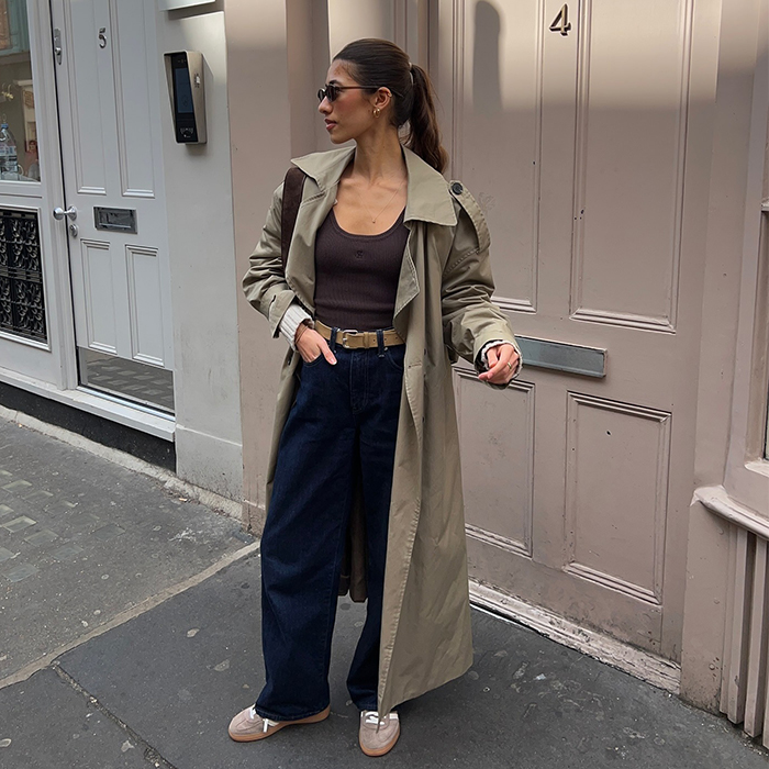 Maya wears black trousers, brown tank, trench coat, and tan adidas. 