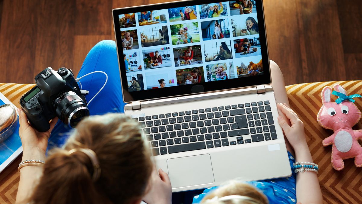 mother and child sitting on sofa in the modern living room viewing image library while transferring photos from DSLR photo camera on a laptop.