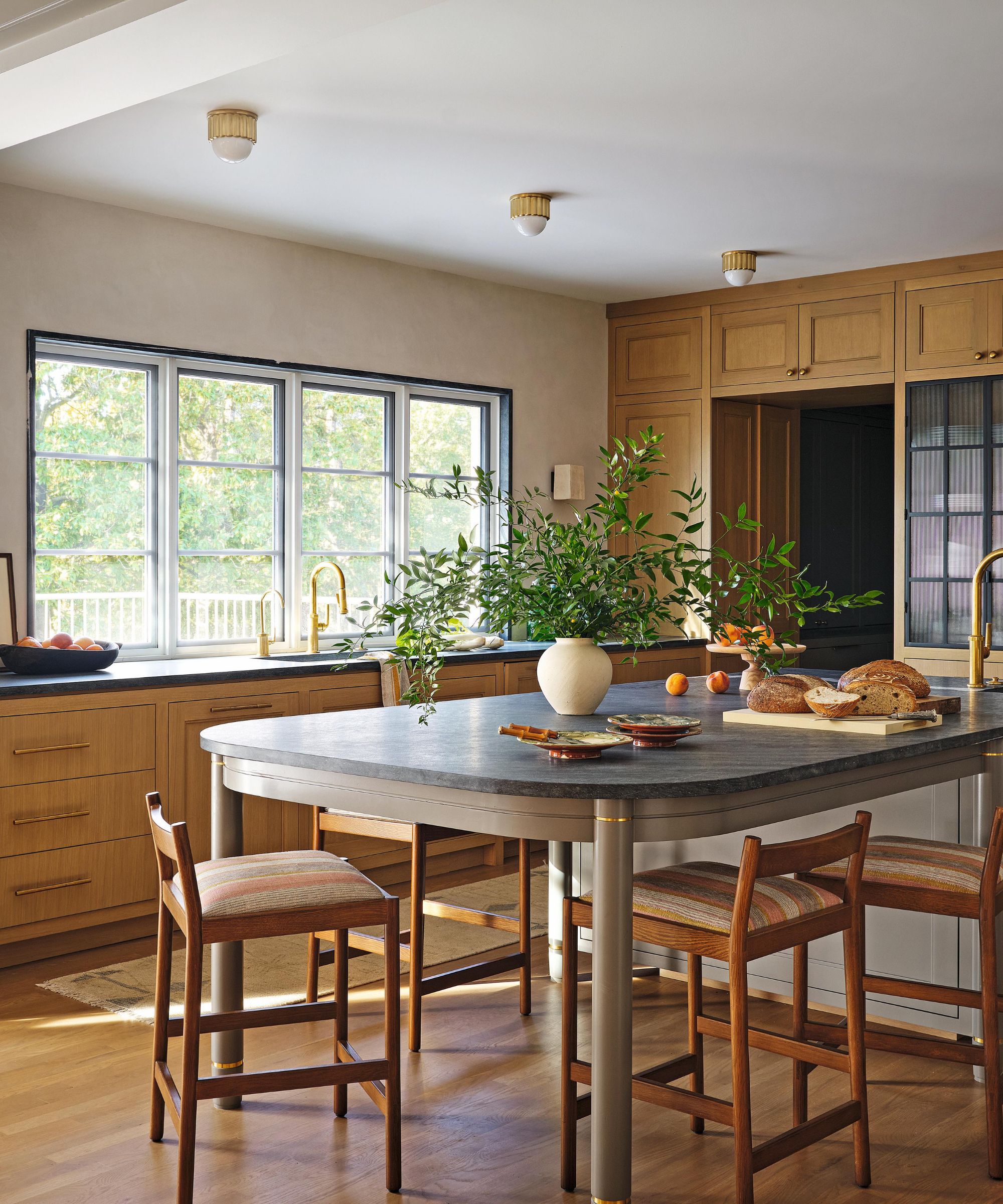 a neutral and oak kitchen with large windows, and a curved table-style kitchen island with bar stools and dark grey stone counters