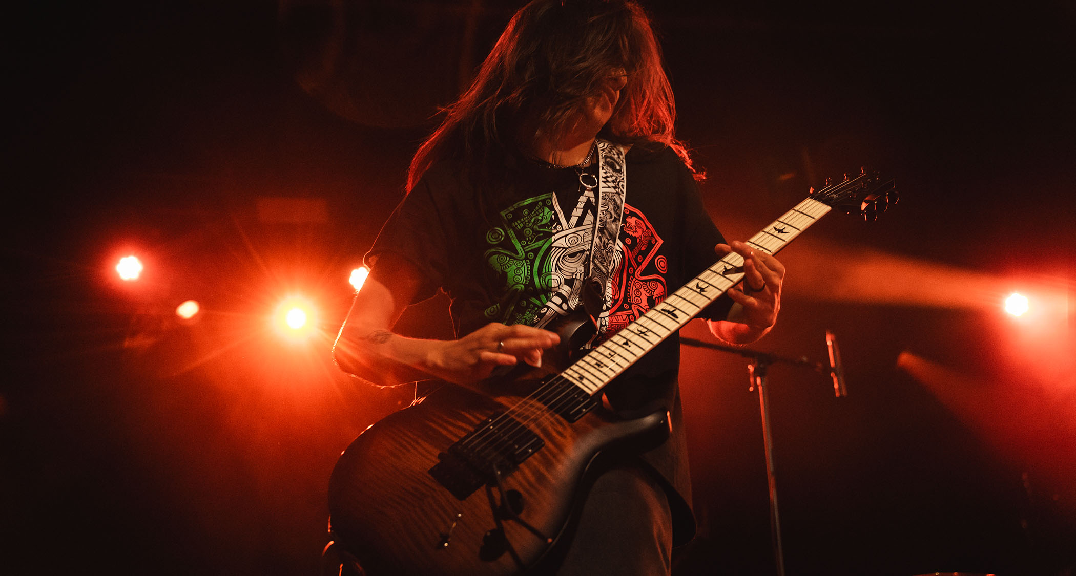 Sebastian Gonzalez riffs on a figured-topped PRS electric guitar, with red lights illuminating the stage.