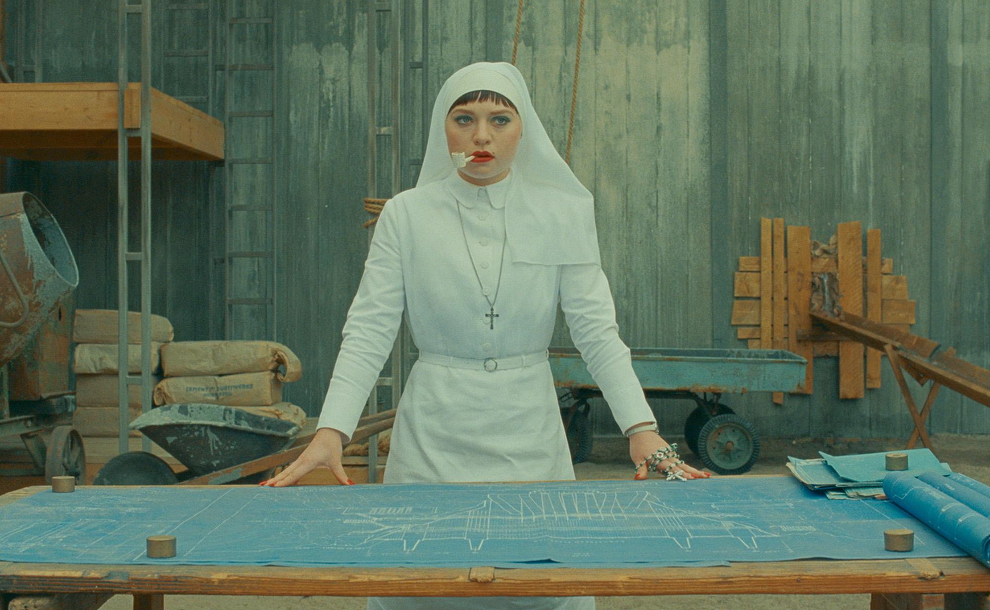 Still from the film The Phoenician Scheme. Mia Threapleton is dressed up as her character Liesl in an all-white nun costume smoking a white pipe. She is standing in front of table with building plans on it.