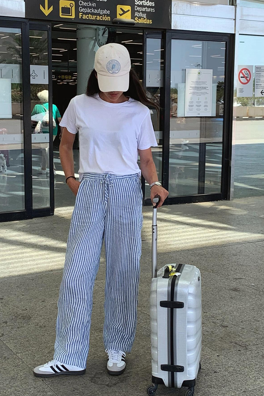 summer airport outfit is shown in a photo of a French woman standing outside the terminal with her white suitcase wearing a white baseball cap, a white t-shirt, striped trousers, Adidas Samba sneakers, and a silver watch