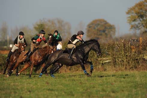 Four horse and rider combinations make a quick turn after jumping a hedge on a team chasing course