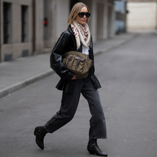 Photo of a woman wearing black jeans, black ankle boots, black jacket, white t-shirt, and sunglasses holding an olive bag.