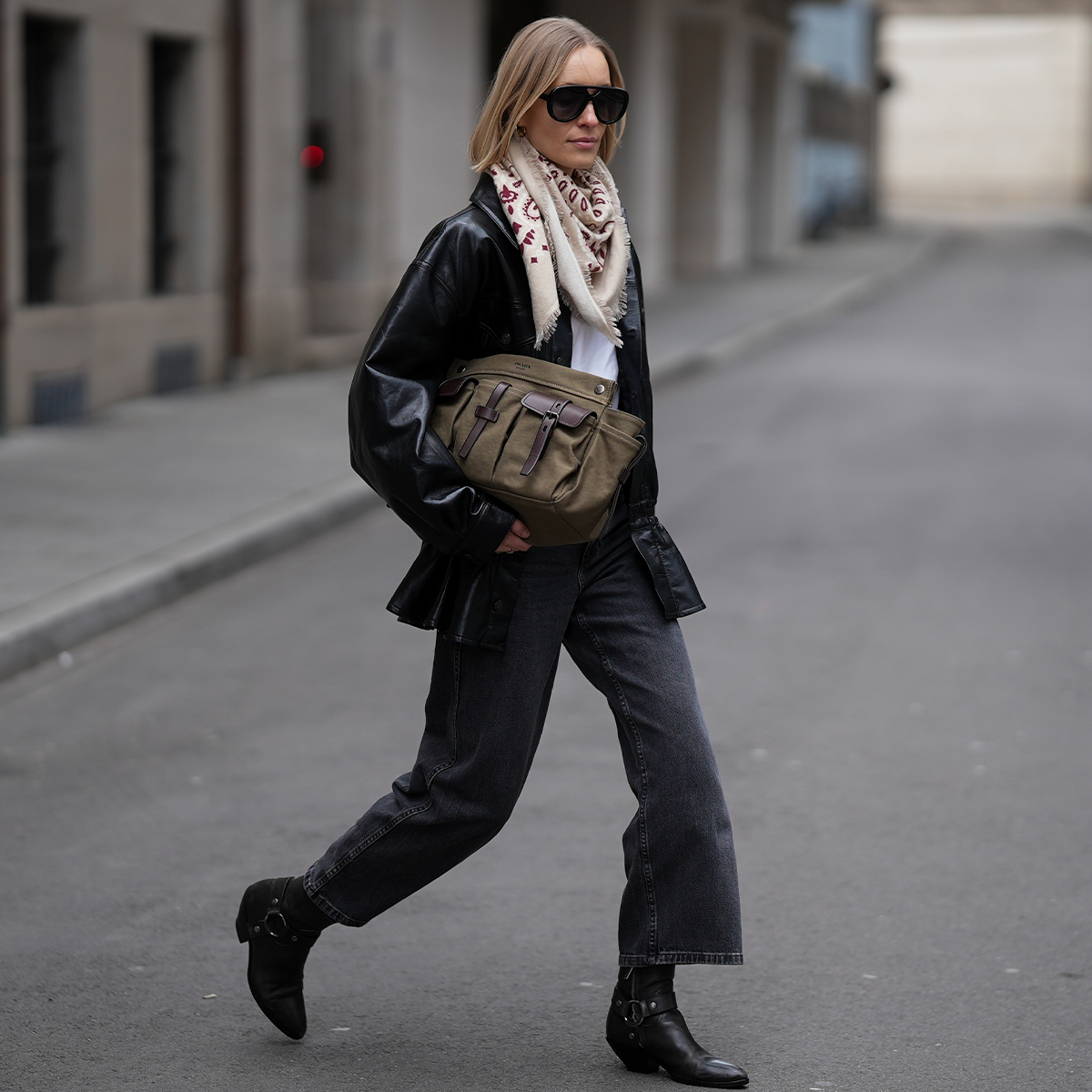 Photo of a woman wearing black jeans, black ankle boots, black jacket, white t-shirt, and sunglasses holding an olive bag.