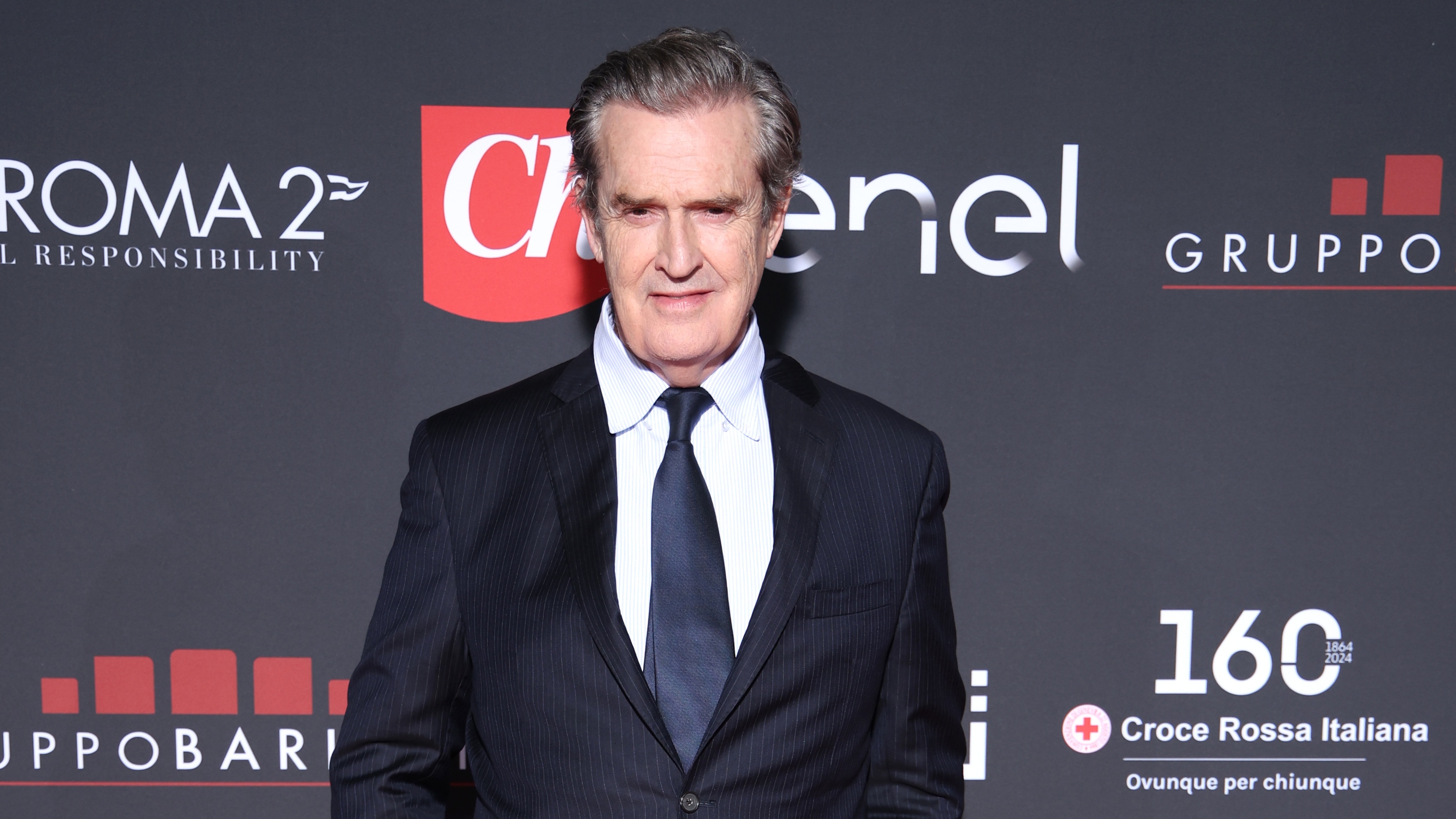 Rupert Everett attends the Red Cross Ball In Rome on November 06, 2024 in Rome, Italy in a dark suit, white shirt and navy tie