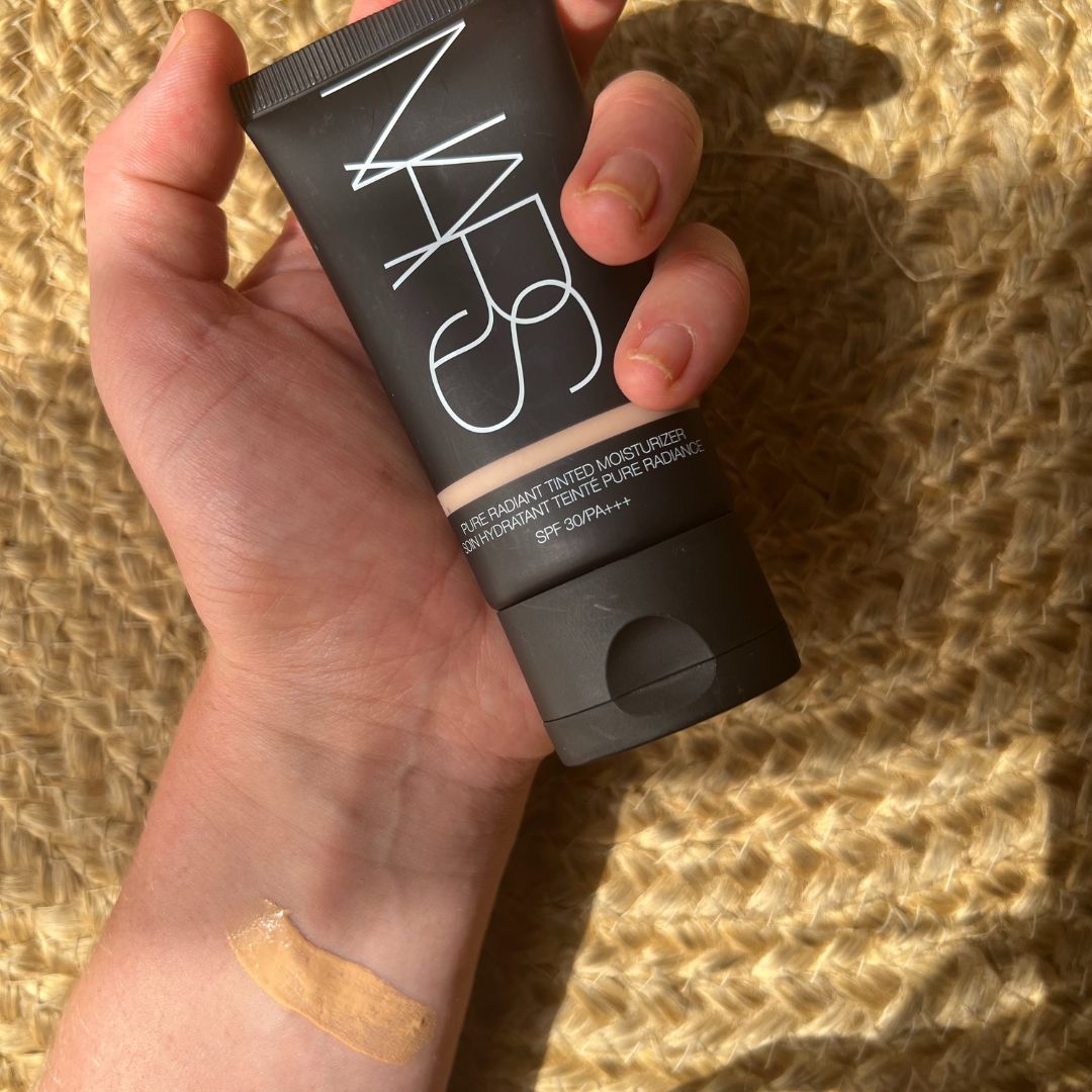 Beauty Editor Fiona McKim's hand holding a tube of foundation, with a swatch of the foundation on her wrist, on a woven jute rug backdrop