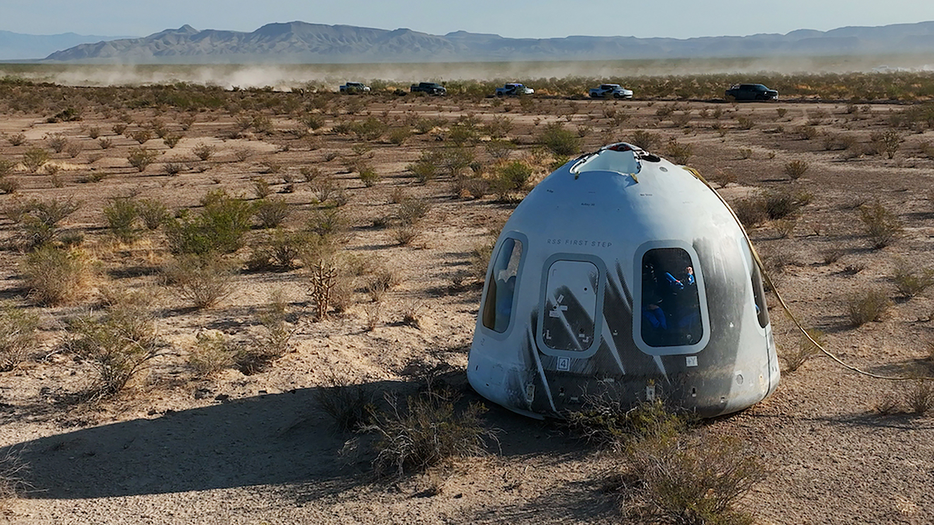 a white space capsule sits in a scrubby desert landscape with mountains in the background