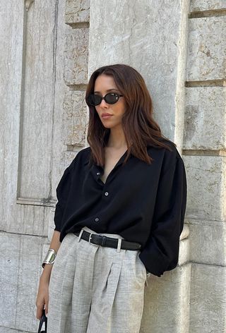 a photo showing the best summer-to-fall transition items with a woman leaning against a wall wearing a tailored black button-down shirt tucked-into brown linen trousers styled with a black belt, silver cuff bracelet, and black sunglasses