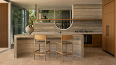 An earthy kitchen with a curved stone island, a floating wood shelf, a vase of flowers, and wood cabinets