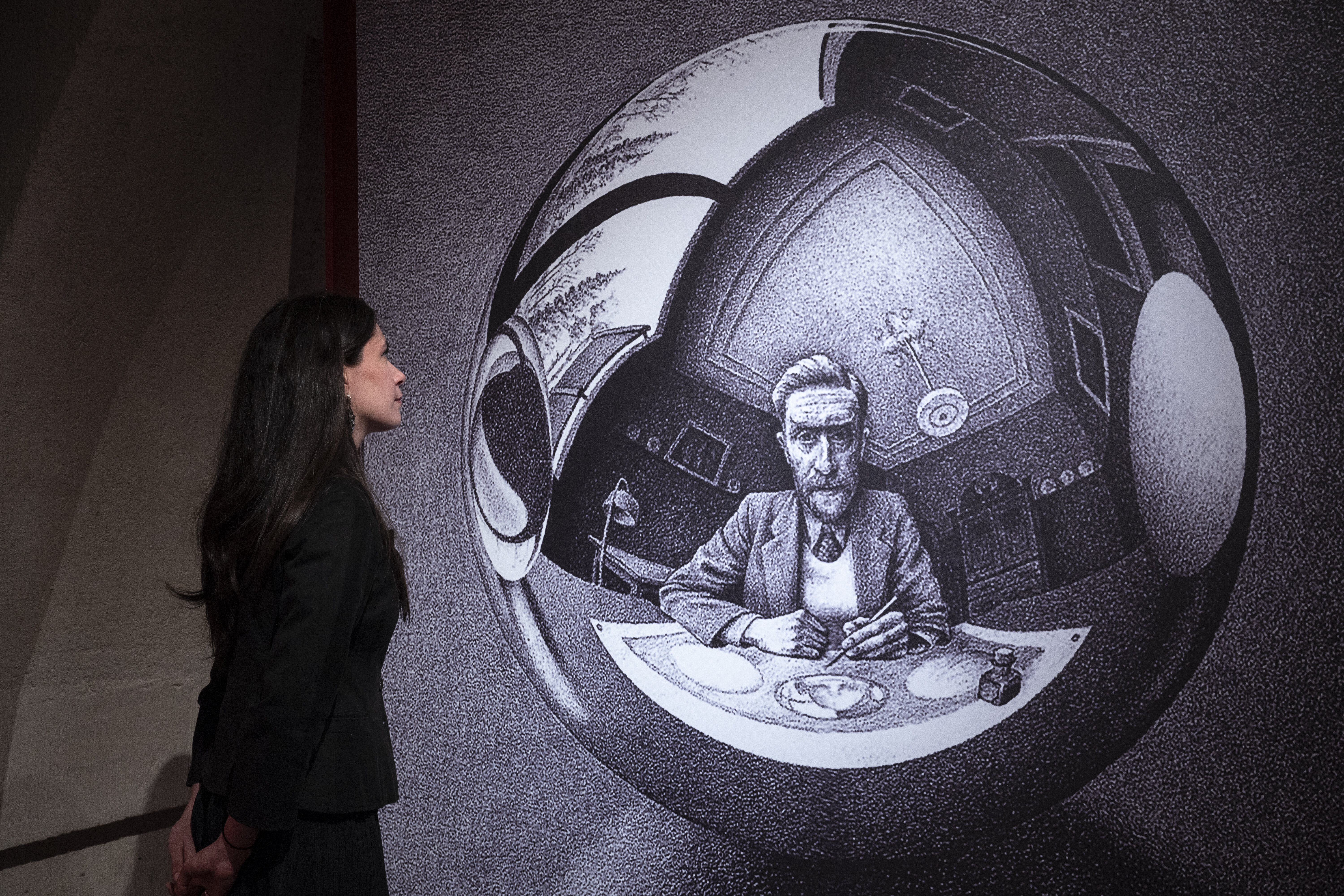 A visitor at Somerset House stands before a large-scale projection of Escher's Hand with Reflecting Sphere, the artist's self-portrait in the curved mirror looming dramatically behind her on the gallery wall