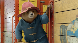 Paddington the bear leaning out of a yellow structure wearing a red hat and a blue coat.