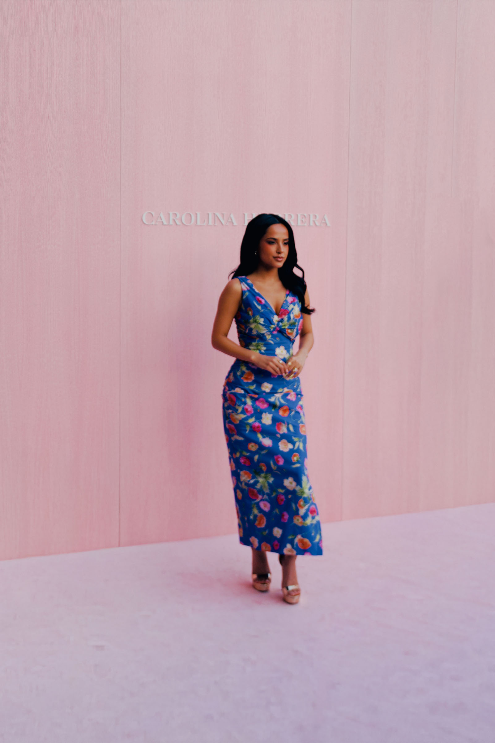 Becky G poses at the Carolina Herrera spring 2026 show in Madrid. She is wearing a cornflower blue sleeveless dress with floral print and metallic heels.