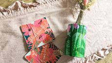 We explain how to decoupage. Here are two coral floral decoupage coasters and a green floral decoupage tin with dried flowers in it, on top of a pink frilled table runner on top of a light wooden table