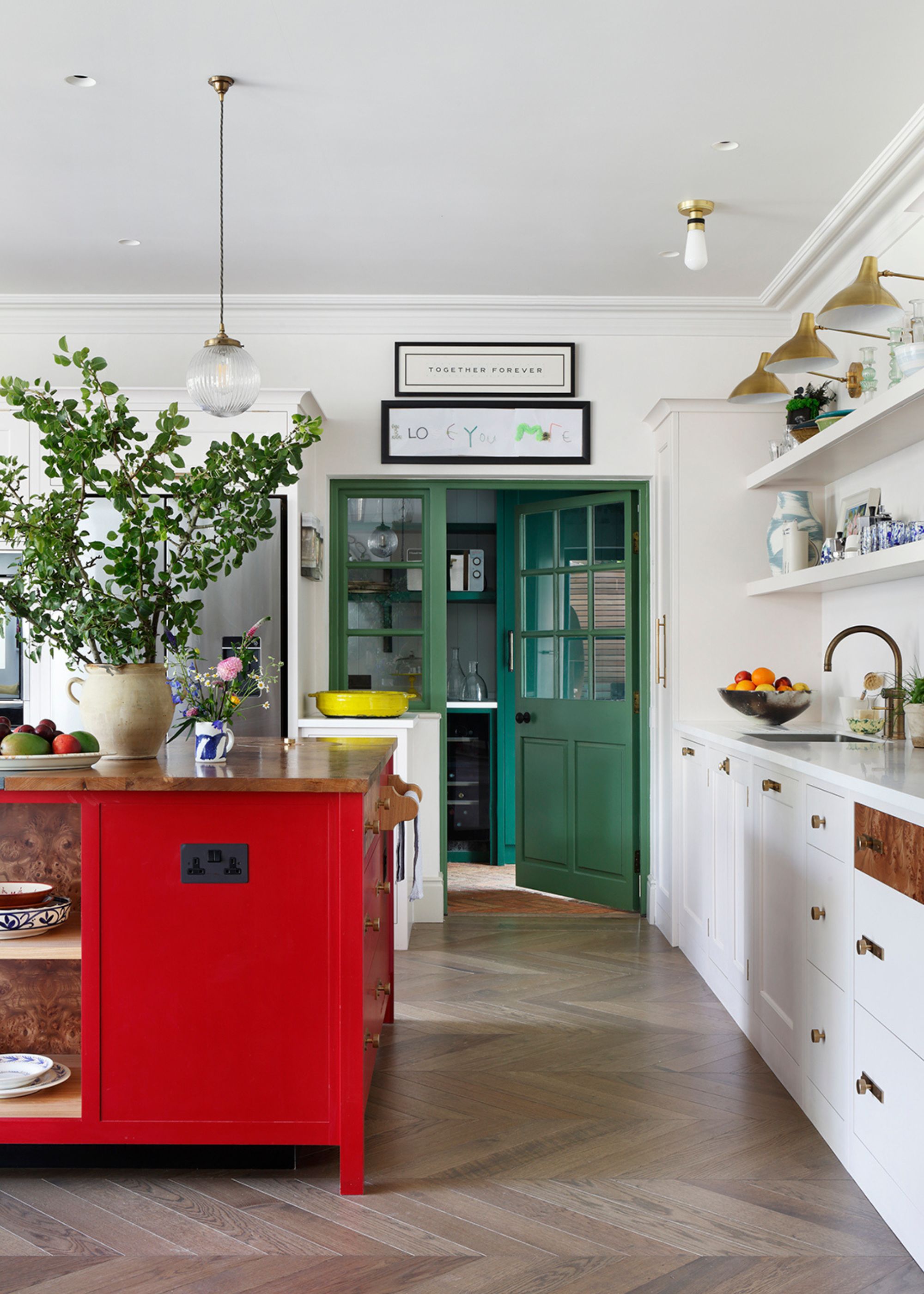 A large kitchen with white walls and cabinets, parquet flooring, a bright red kitchen island with a large vase of foliage, and dark green French doors leading into a pantry.