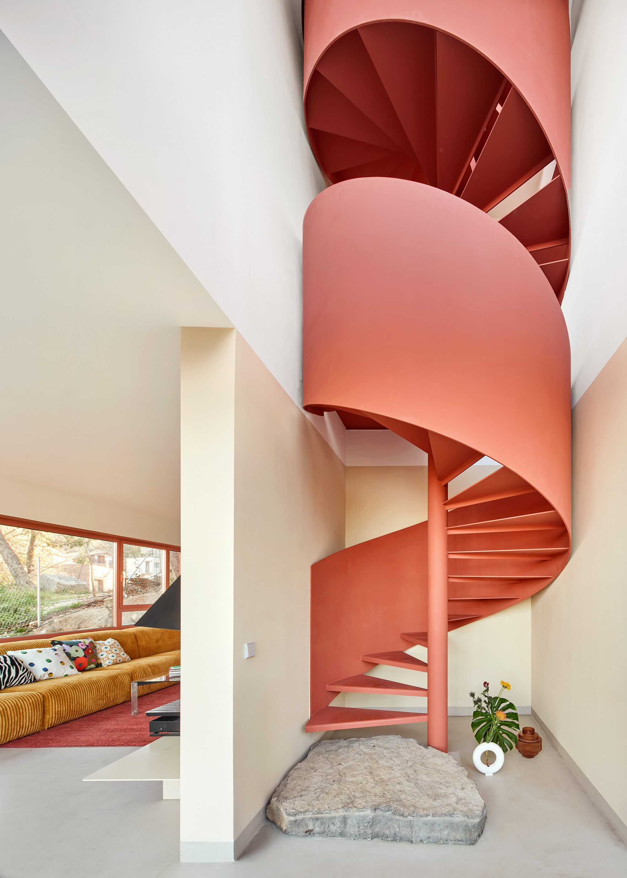 A modern, terracotta colored spiral staircase with a few into a living room with a mustard corduroy sofa
