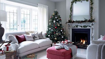 Christmas living room, faux Christmas tree with red and god baubles, lit open fireplace, chimney breast, mirror with fairy light garland, cream sofa, board games, pink ottoman, leaded French doors.