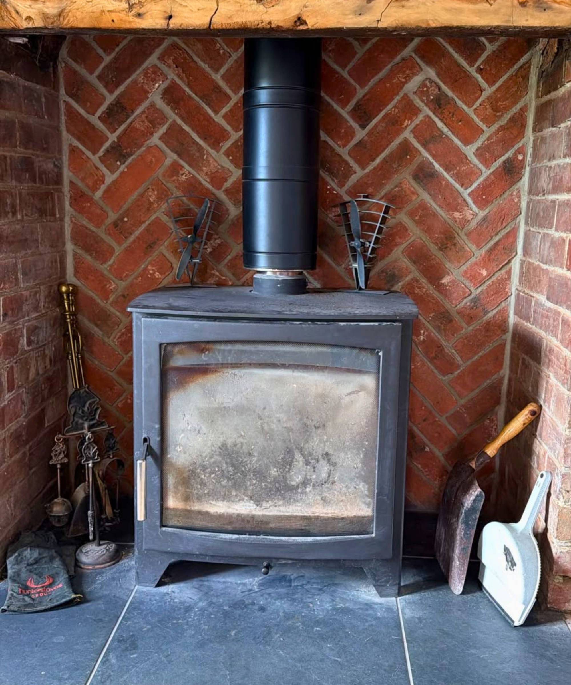 Wood burner with sooty glass door and heat-powered fans on top, set in a herringbone brick fireplace.