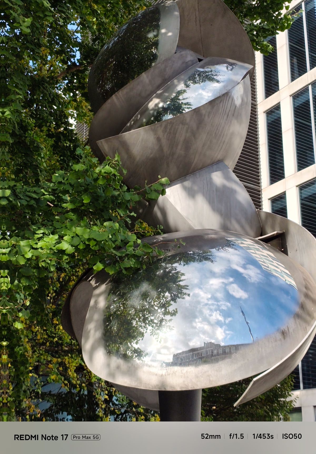 Abstract polished metal sculpture surrounded by trees and modern buildings.
