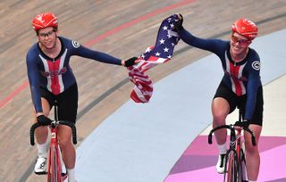 two women on a cycling track, on bicycles, holding an american flag