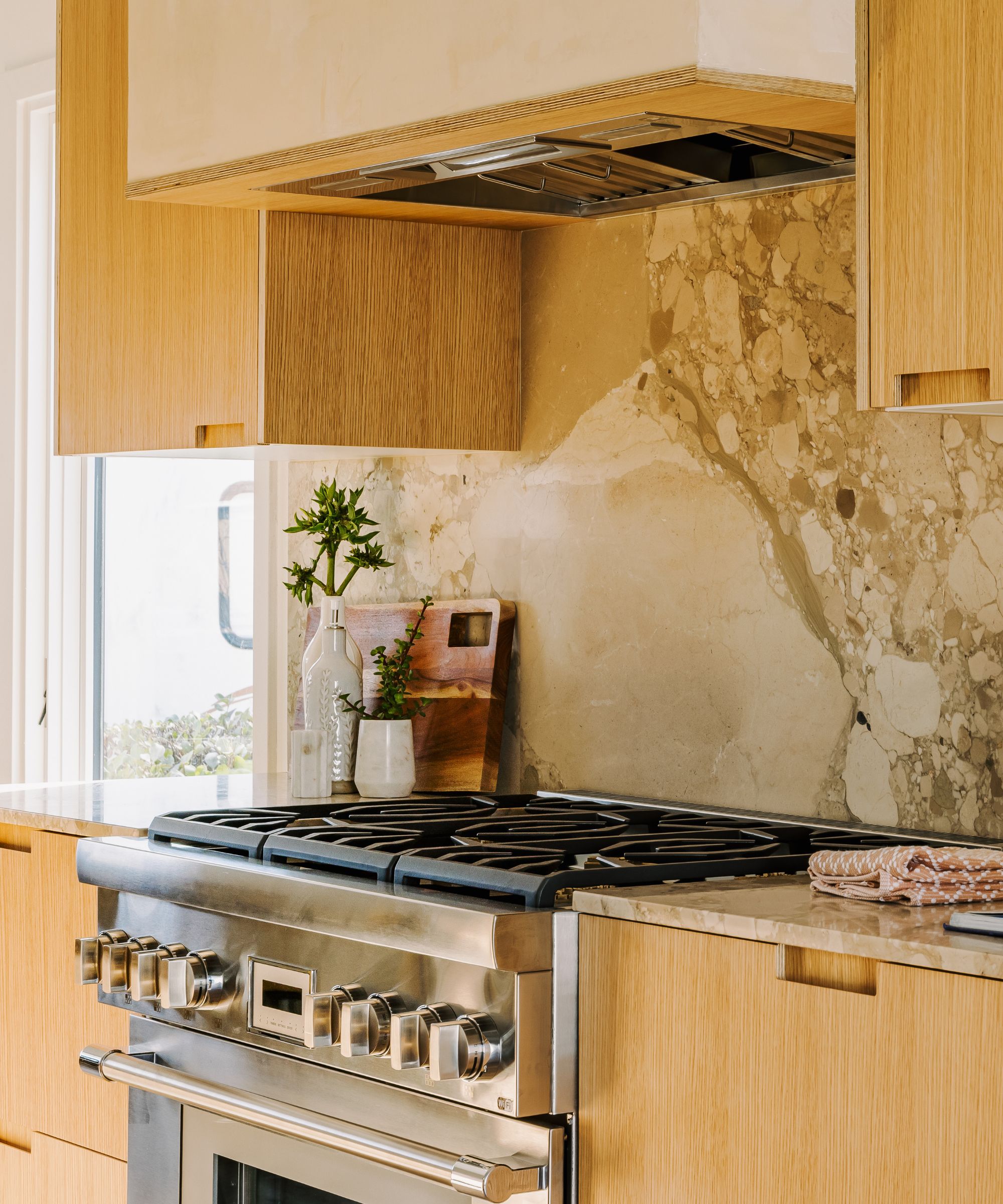 Detail shot of a high-end stainless steel gas range integrated into light wood cabinetry with a dramatic, large-scale stone slab backsplash.