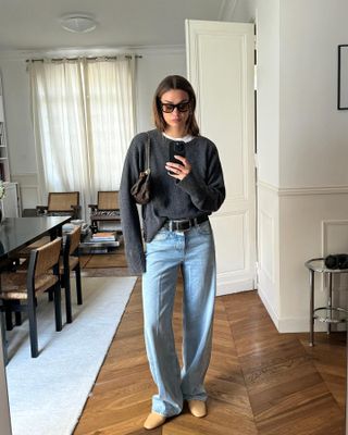 French fashion influencer Anne-Laure Mais poses for a mirror selfie in her Paris apartment wearing sunglasses, a charcoal gray crewneck sweater layered over a white tee, mini Louis Vuitton chain shoulder bag, black belt, relaxed jeans, and neutral flats.