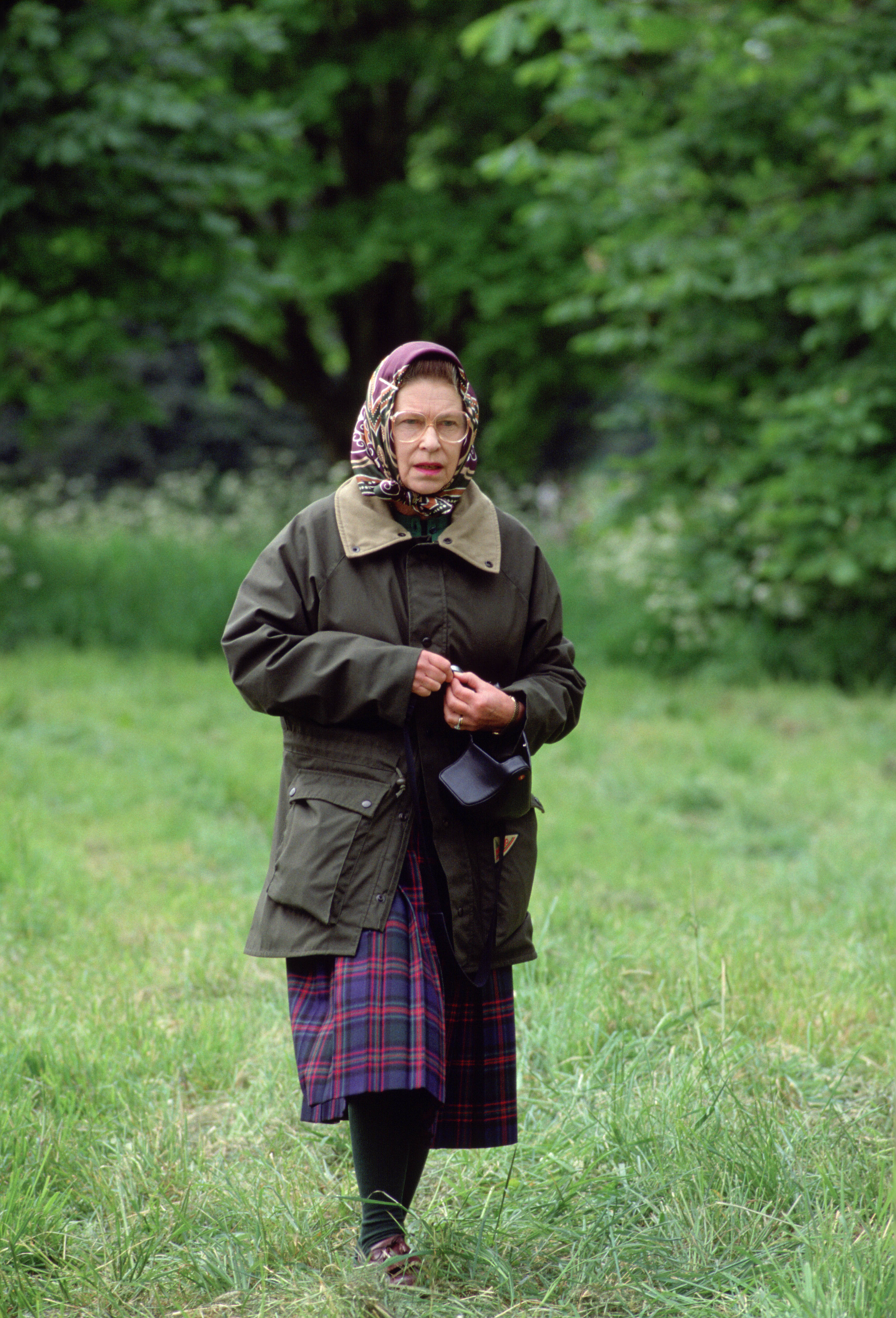 Elizabeth II in a wax jacket and tartan skirt striding through grass