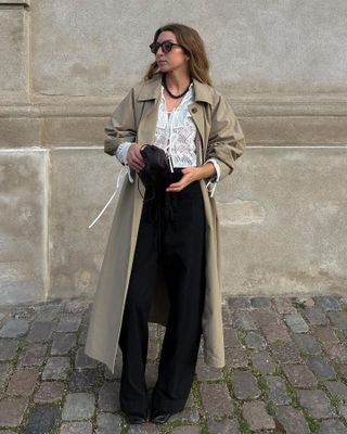 British fashion influencer Brittany Bathgate poses on a European cobblestone street in a trench coat outfit with a beaded necklace, tie front white top, black pants, and boots