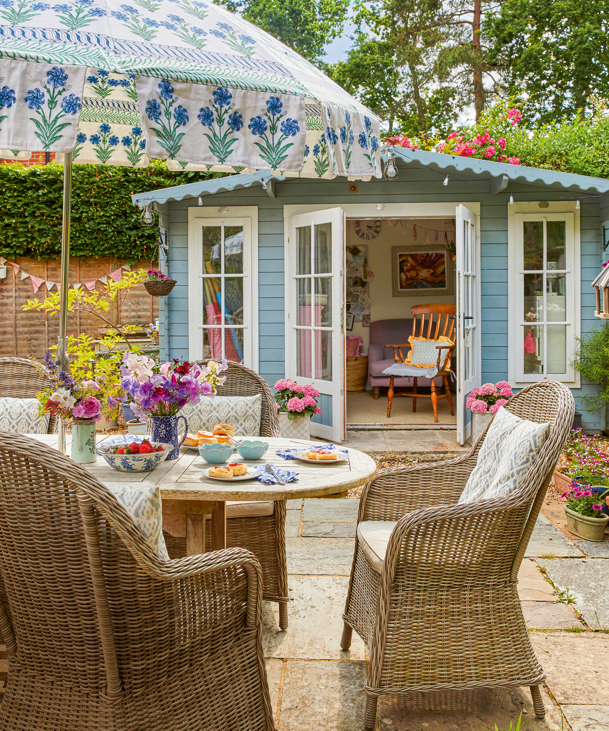 outdoor patio with rattan dining chairs, patterned parasol and blue painted summerhouse behind