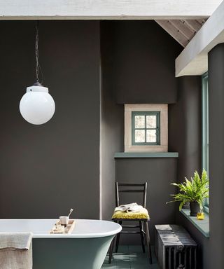Dark gray painted bathroom with green flooring and matching bathtub, light wood ceiling beams, rounded white glass pendant light, small window, black chair below window