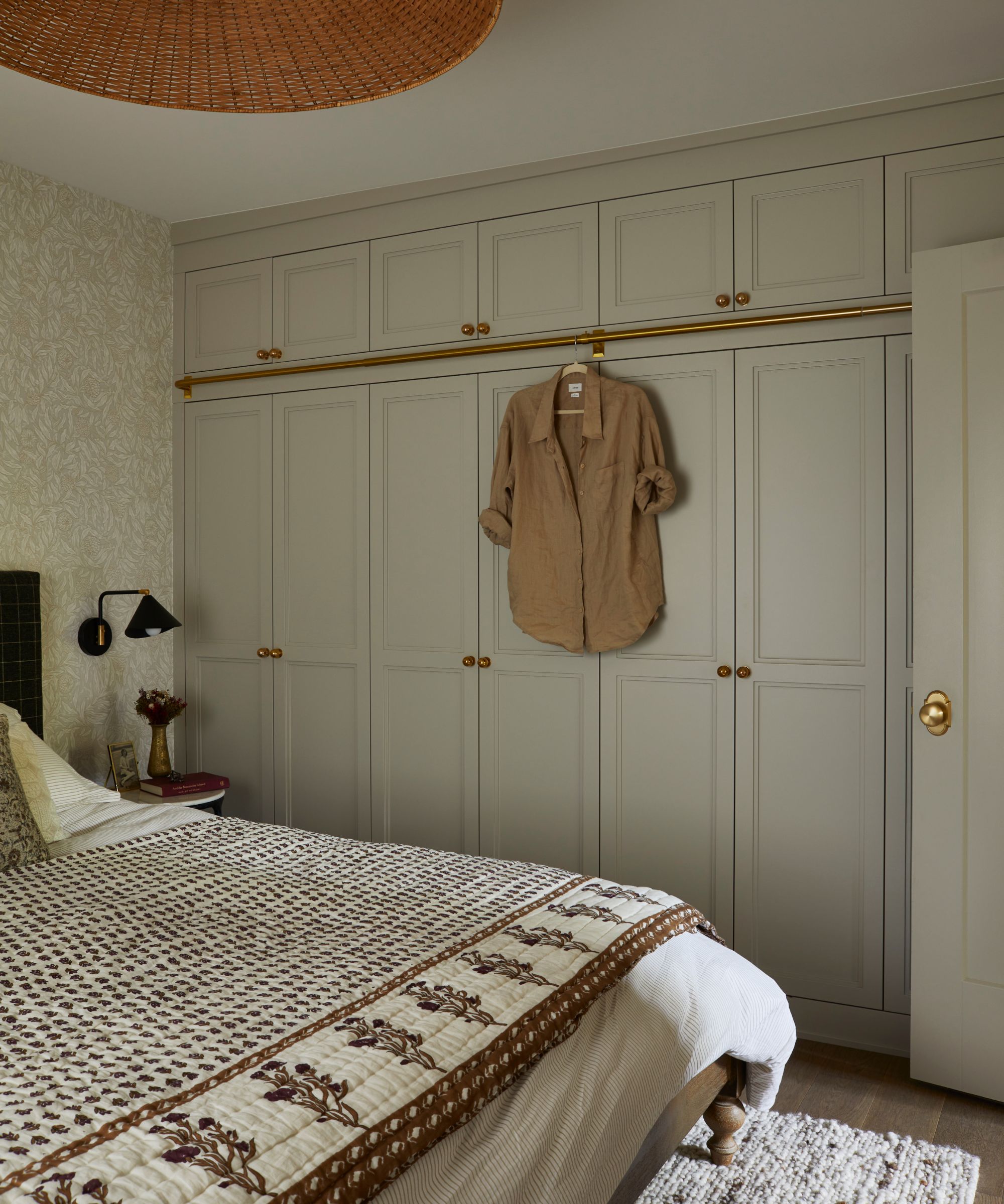 a calm primary bedroom with neutral floral wallpaper, custom wardrobes, and a bed with a large floral bedspread