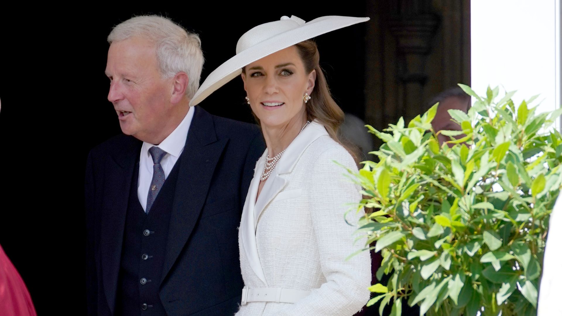 Catherine, Princess of Wales emerges from Windsor Castle wearing a white lace-trim skirt with a tonal jacket neatly fastened around her waist. She wear a wide-brim white hat, dark beige heels and carries a small satin clutch in her hands.