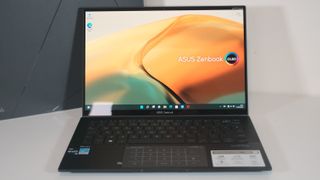 An ASUS Zenbook laptop with a dark chassis, thin bezels, and an OLED screen displaying a colorful abstract wallpaper, set on a white surface.