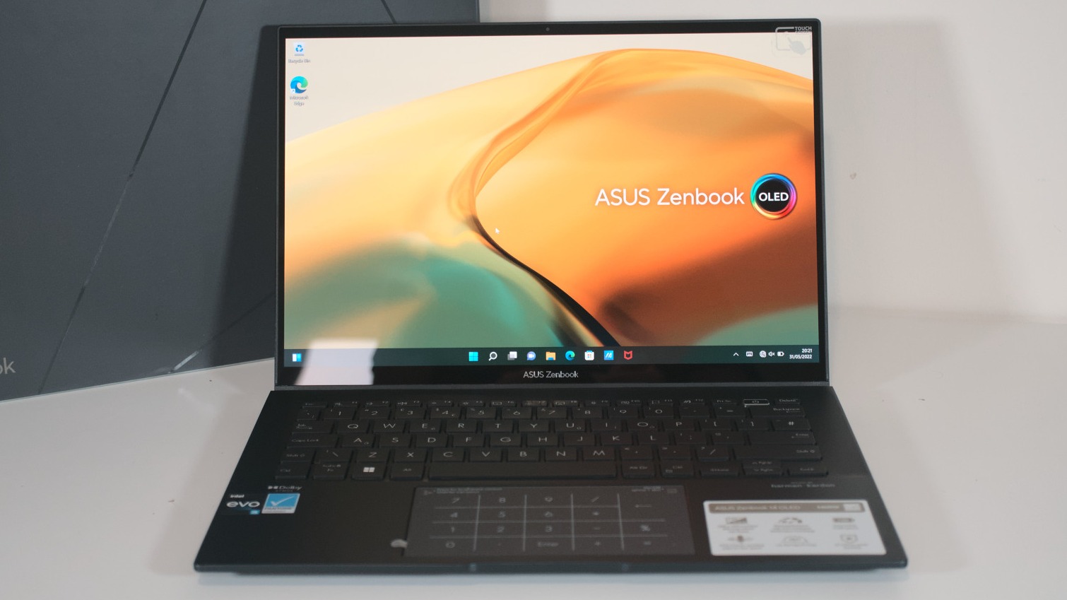 An ASUS Zenbook laptop with a dark chassis, thin bezels, and an OLED screen displaying a colorful abstract wallpaper, set on a white surface.
