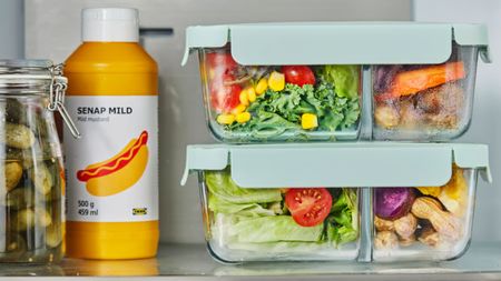 A stack of two glass bento food boxes with two compartments and blue lids on top of one another, filled with salad and vegetables, on a fridge shelf beside a bottle of yellow mustard. 