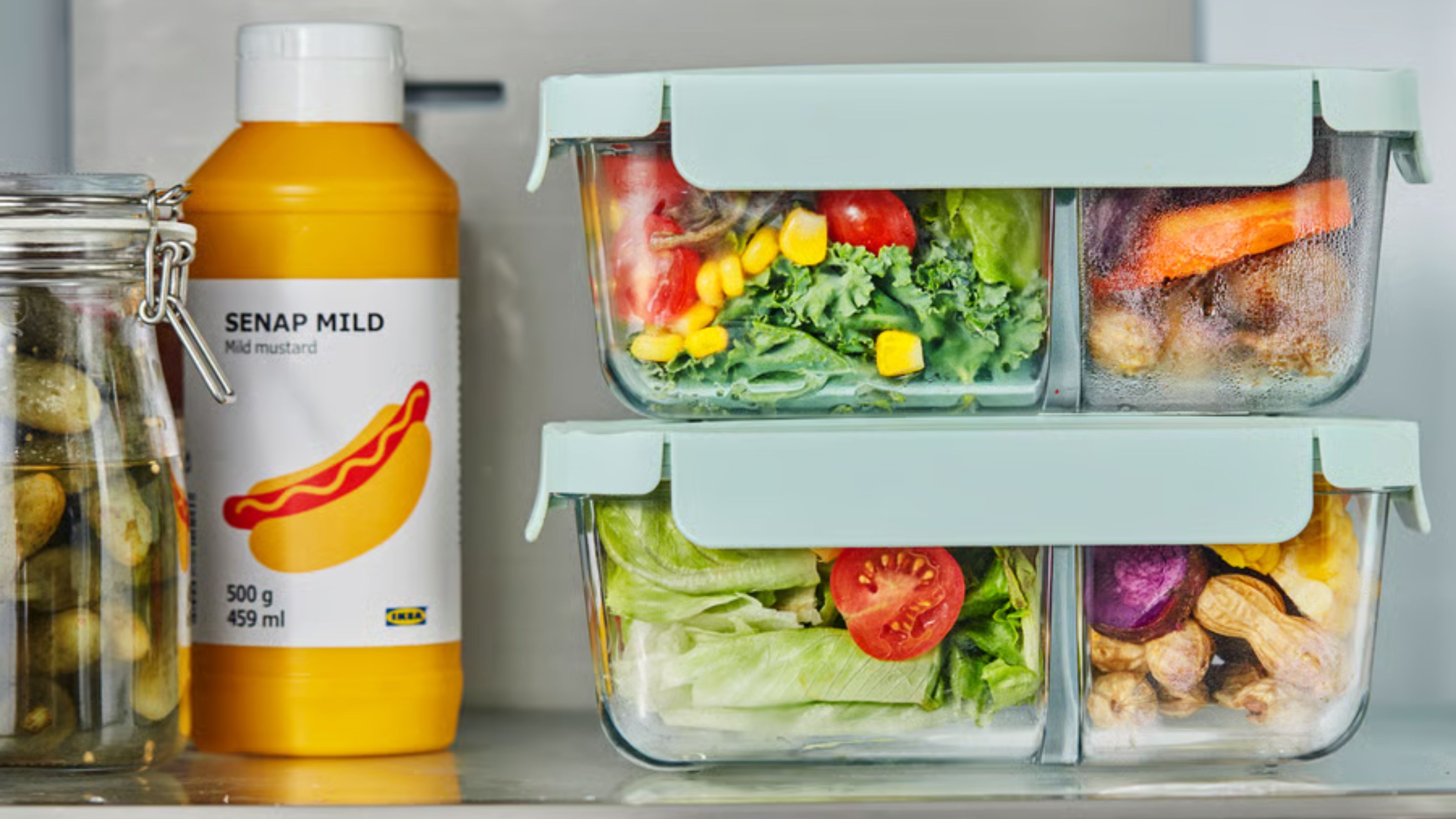 A stack of two glass bento food boxes with two compartments and blue lids on top of one another, filled with salad and vegetables, on a fridge shelf beside a bottle of yellow mustard. 