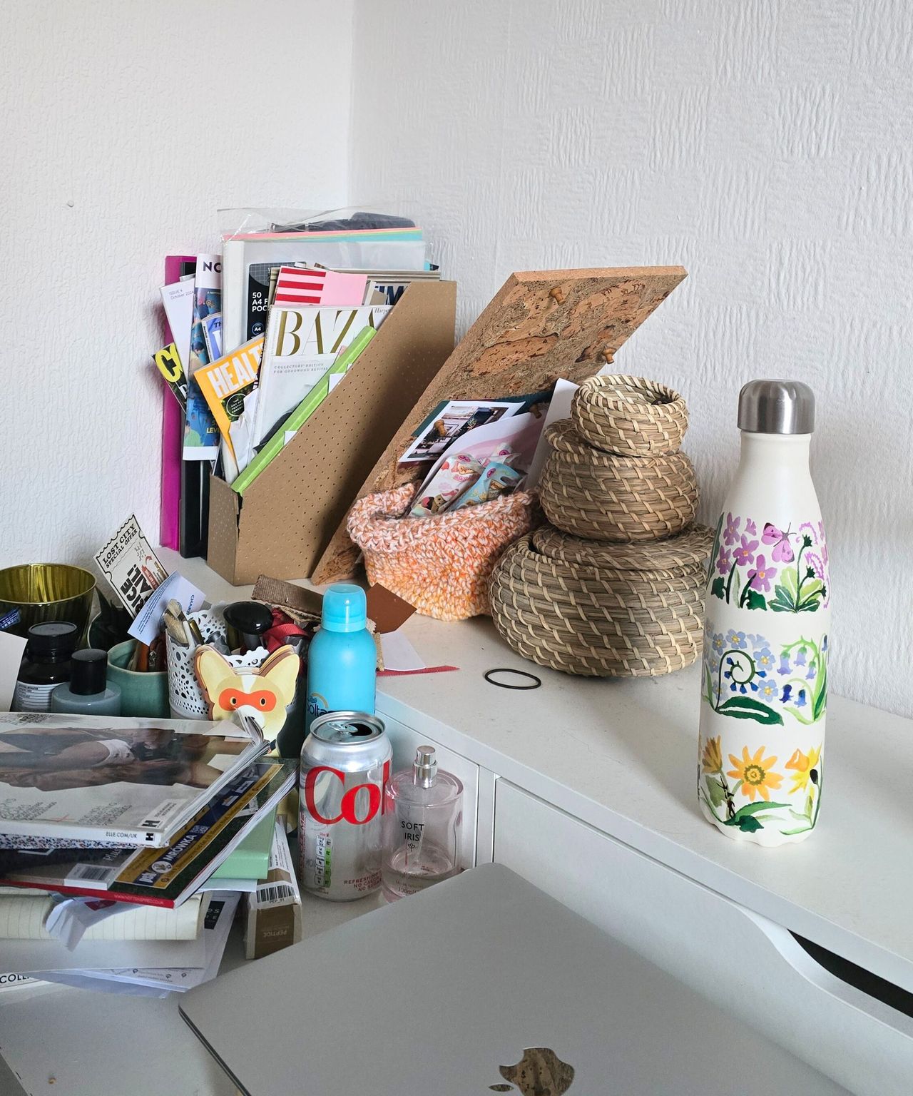 A white desk with a water bottle, woven circular boxes, a full magazine filer, a pile of magazines, and a silver laptop on top of it