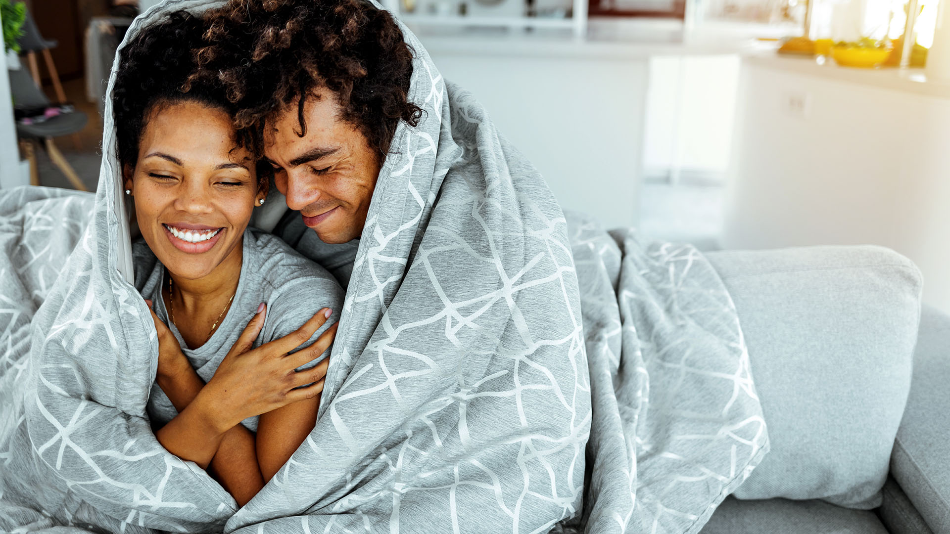 A couple sit cuddling on a bed wrapped in a blanket