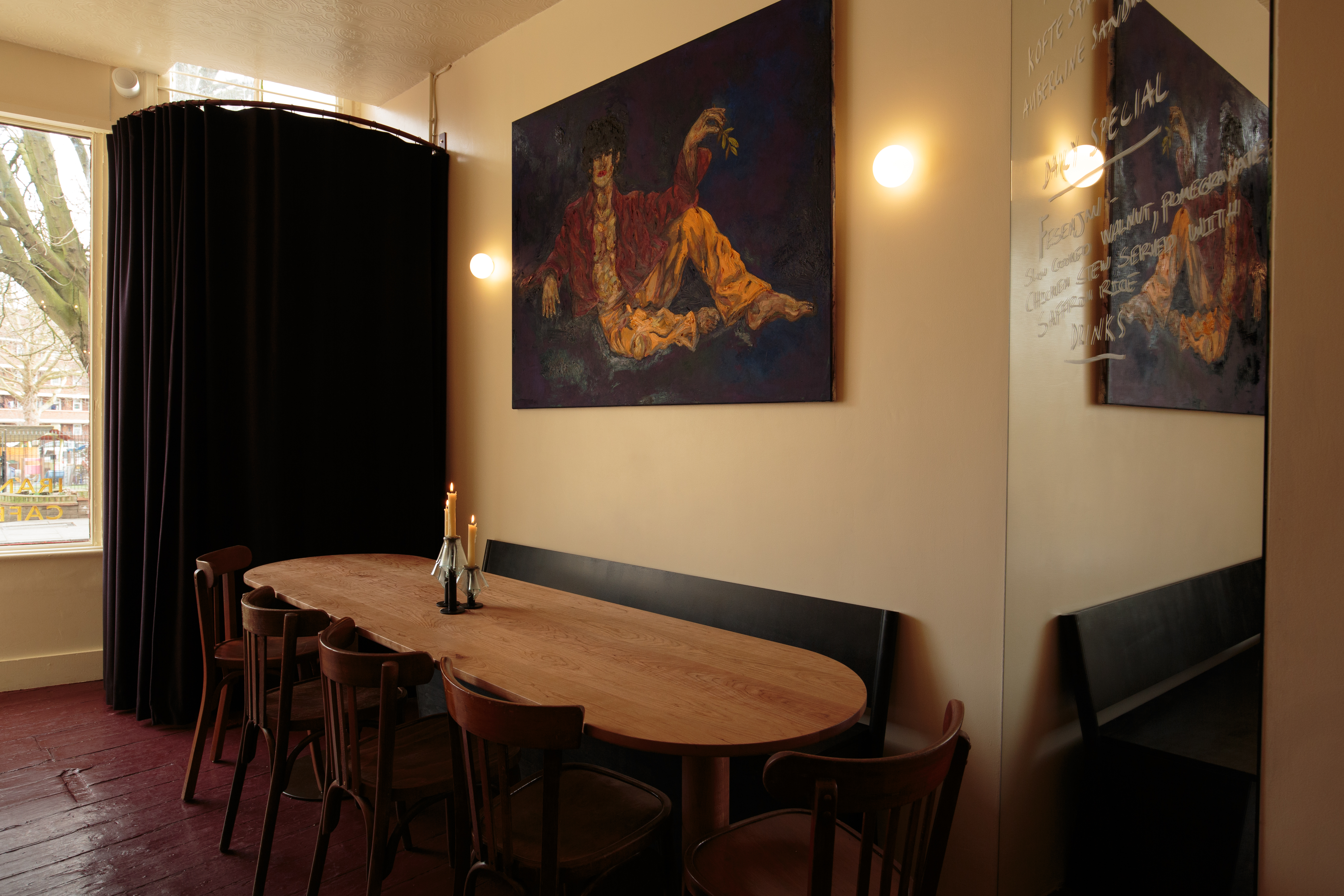 A dining room with red-painted floors, a semicircular entrance curtain in dark blue, figurative paintings hung on beige walls, and an oblong wooden tables with red chairs and a black banquette.