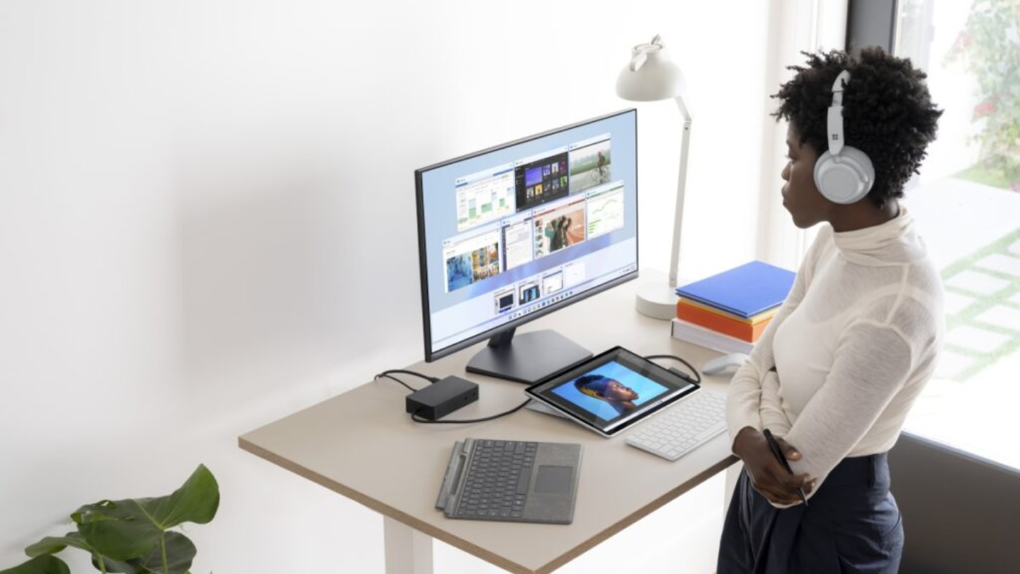 A desk with a lamp, moniter, tablet and two keyboards on it with a woman looking at it wearing white earphones