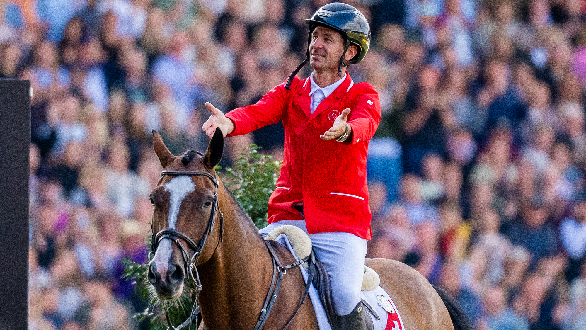 Steve Guerdat, a Swiss rider in a red coat, holds out his hands to praise his horse, Dynamix De Belheme, a bay mare with a white blaze, after they become 2026 world showjumping champions in Aachen
