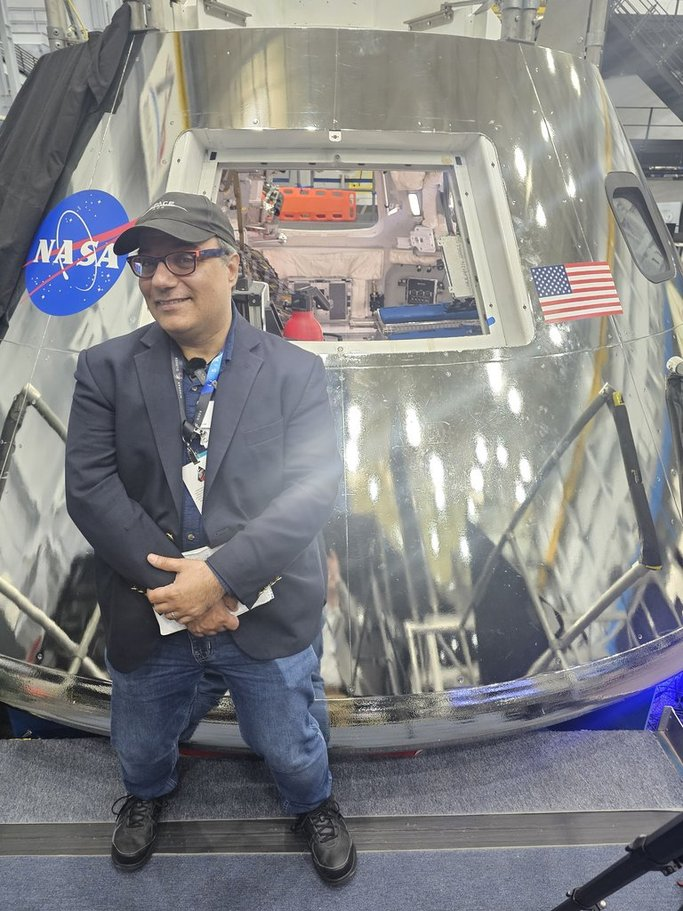 A man in a suit jacket stands in front of a metallic spacecraft capsule branded with the NASA logo.