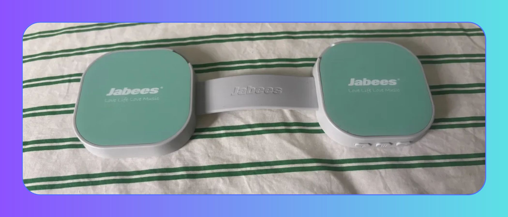 The image shows a flat dumbbell-shaped green and white under pillow speaker, placed on green and white striped bed sheets. This image is set against an ombre blue background 