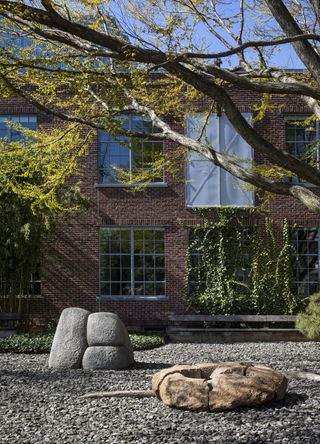 An urban garden features organically shaped sculptures in rock and wood, green-leafed trees, and a red brick building with wide loft-style windows.