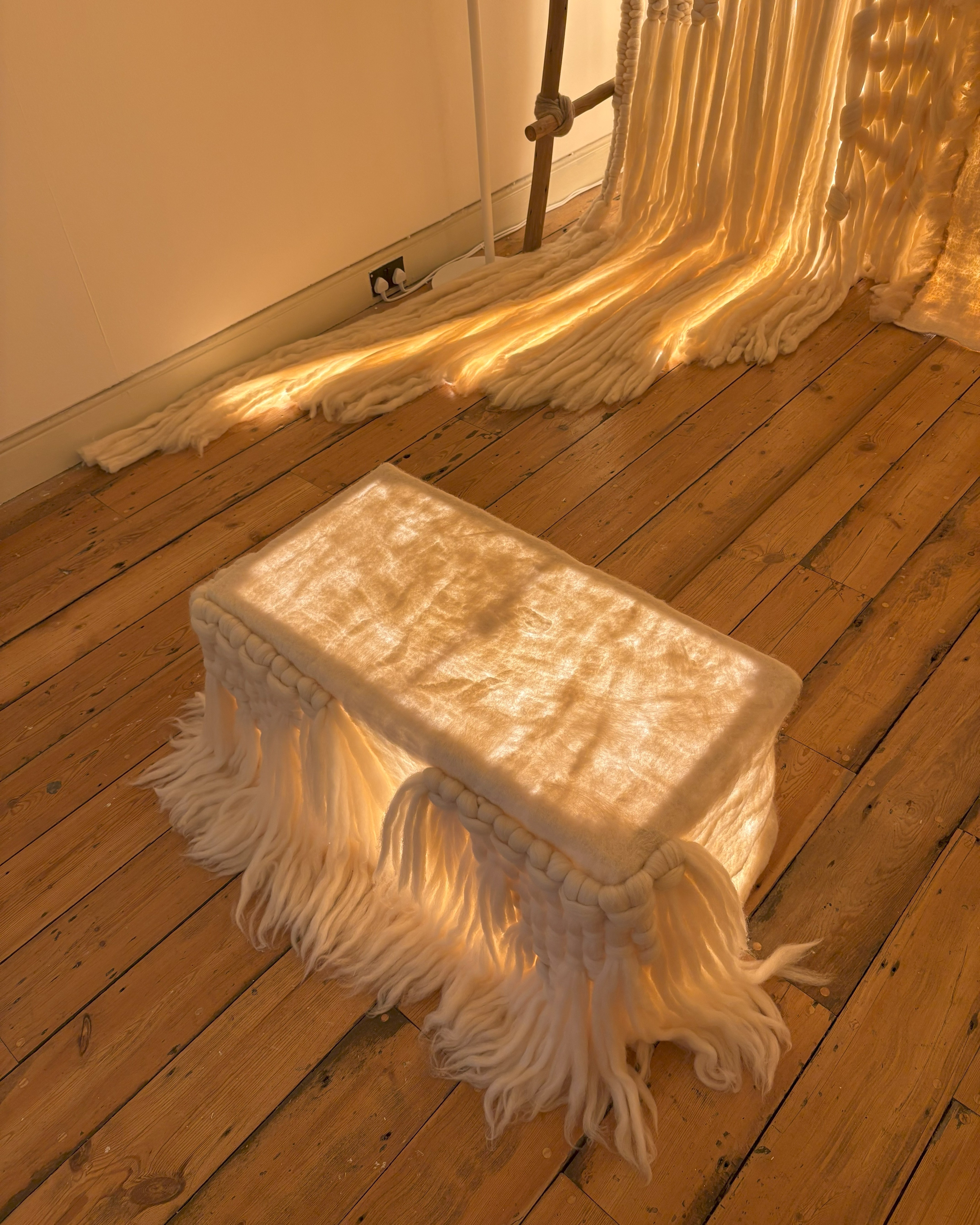 A back-lit textile installation features a handwoven Ottoman sitting in front of a large-scale, strikingly woven panel in a wooden-floored room.